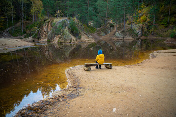 The child is resting on a bench at the edge of the lake in the middle of the forest