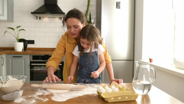 Tenderly portrait of mother and little cute daughter cooking together, lovely mom kneads the dough, adorable toddler girl helping her, they looking at each other, spends leisure at home,baking cookies