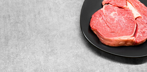 High angle studio shot of a raw beef rib eye steak on a black design plate standing on a grey concrete surface, with free copy space. Keto diet, warrior diet concept. Raw red meat ready for barbecue.