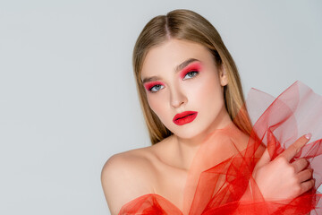 Fair haired woman with red makeup holding tulle and looking at camera isolated on grey.