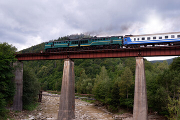 An old diesel train with passenger cars moves along the bridge over the mountain slopes above the seething river. Ukraine. Carpathian mountains.