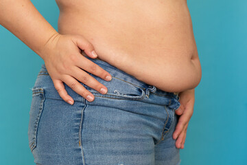 Side view of unrecognizable fat plump obese overweight woman stand in blue jeans putting hand on waist, showing excess naked belly on blue background. Body positive, obesity, weight loss, liposuction.