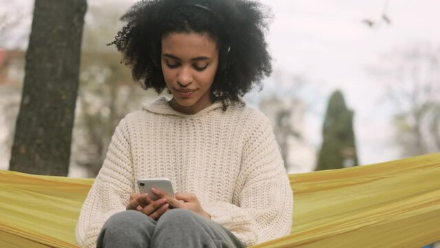 Attractive curly African American woman in headphones sitting in a yellow hammock, listening music and scrolling her social media. High quality 4k footage