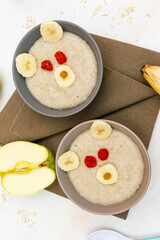 Funny cute kids childrens baby's healthy breakfast lunch oatmeal porridge in bowl look like bear face decorated with apple, banana, dried berry fruits. dessert food art on white wooden table