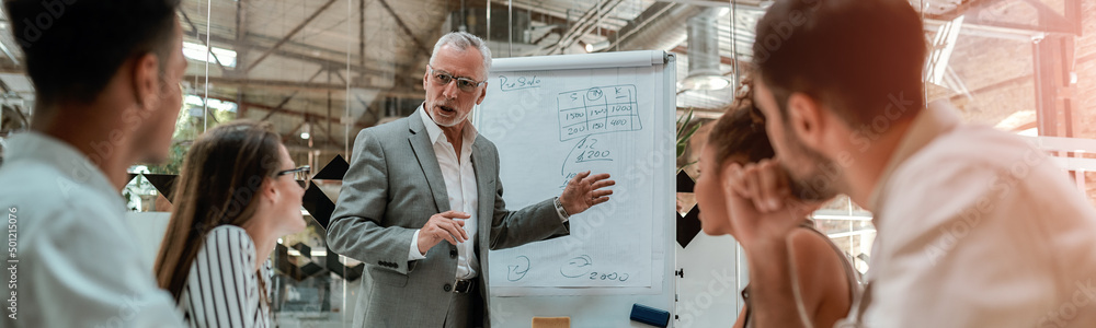 Wall mural confident mature businessman pointing at white blackboard and explaining something to his young coll