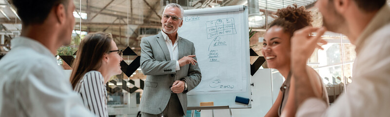 New business strategy. Mature businessman standing near whiteboard and explaining something to his...