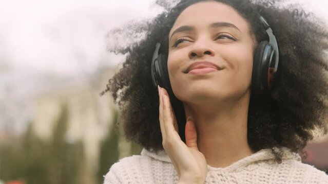 Attractive young African American woman listening to music in headphones standing in the park. Portrait of beautiful curly brunette. High quality 4k footage