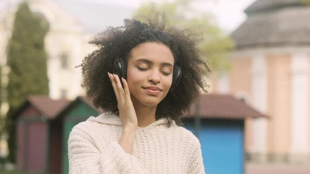 Attractive young African American woman listening to music in headphones standing in the park. Portrait of beautiful curly brunette. High quality 4k footage