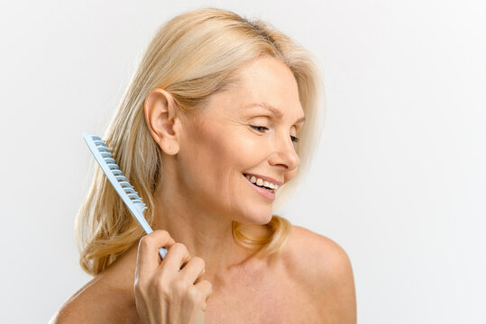 Elegant serene middle-aged blonde woman combing with crest, looks down and laughing cheerfully isolated on white, mature lady caring her hair, everyday routine concept