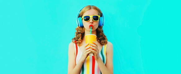 Portrait of stylish young woman listening to music in headphones with fresh cup of juice on blue background