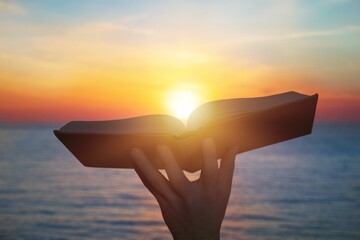 Hand holding open bible book up to the sun light.