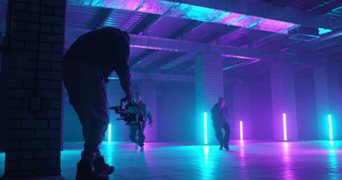 Backstage of video shooting. Cameraman filming hip-hop dancers in neon light. Operator moves with image stabilizer in hands at dark studio. Shooting a clip