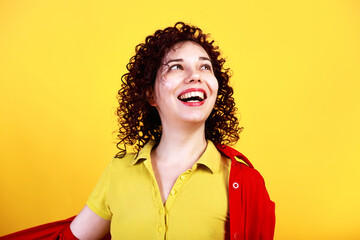 Studio shot of positive young female, looking aside cheerfully and smiling widely. Curly girl in bright shirt and yellow t-shirt. Isolated on yellow background.