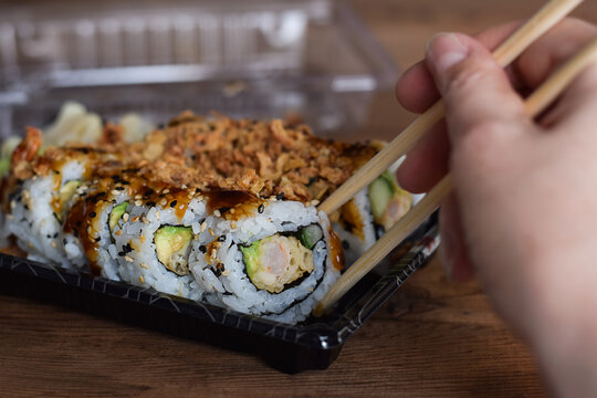 Persons hand taking crispy dragon roll sushi with chopsticks from take out container