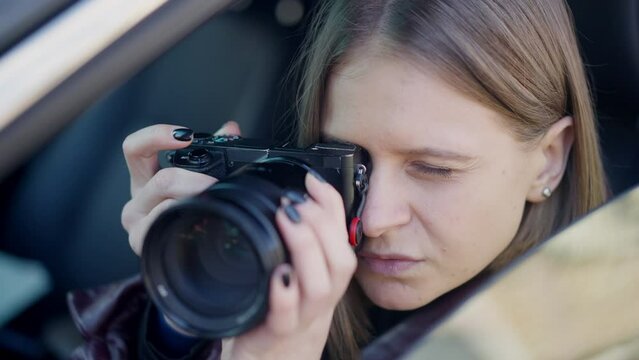 Close-up female spy taking photos with camera sitting in car on driver's seat. Professional serious smart Caucasian woman photographing target outdoors. Evidence and peeping concept