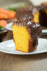 Close up piece of Brazilian carrot cake with chocolate frosting on wooden table with carrots in the background