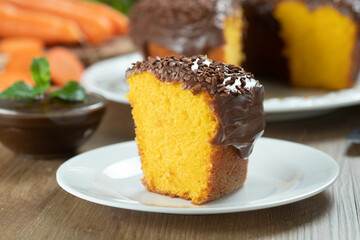 Close up piece of Brazilian carrot cake with chocolate frosting on wooden table with carrots in the background