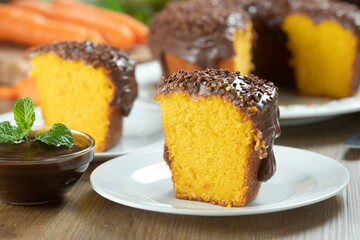 Close up piece of Brazilian carrot cake with chocolate frosting on wooden table with carrots in the background