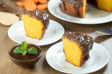 Close up piece of Brazilian carrot cake with chocolate frosting on wooden table with carrots in the background