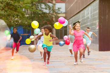Portrait of cheerful preteen girls and boys with colorful toy balloons in hands running on city street on sunny summer day - obrazy, fototapety, plakaty