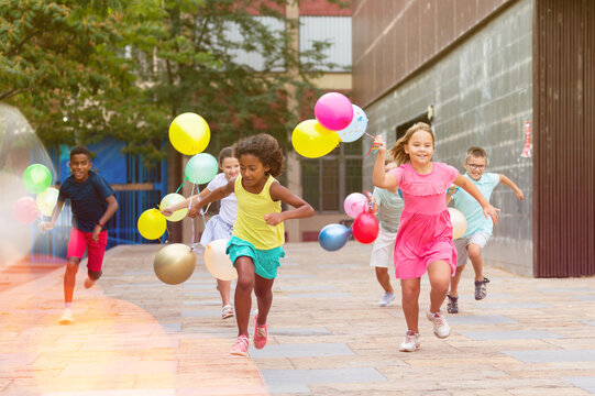 Portrait of cheerful preteen girls and boys with colorful toy balloons in hands running on city street on sunny summer day