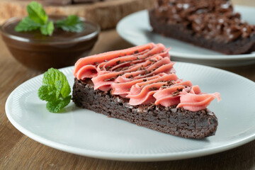 Piece of Chocolate Brownie Slice with Strawberry and Chocolate Icing. Wooden table with mint and chocolate chips in the background