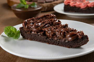 Piece of Chocolate Brownie Slice with Strawberry and Chocolate Icing. Wooden table with mint and chocolate chips in the background