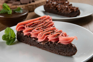 Piece of Chocolate Brownie Slice with Strawberry and Chocolate Icing. Wooden table with mint and chocolate chips in the background