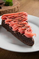 Piece of Chocolate Brownie Slice with Strawberry and Chocolate Icing. Wooden table with mint and chocolate chips in the background