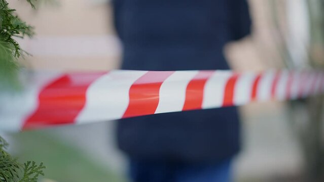 Red and white police tape outdoors with blurred unrecognizable criminalist walking at background. Crime scene with woman investigating murder. Forensics and homicide concept
