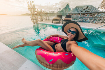 Relaxing woman floating in donut inflatable swimming pool toy at luxury resort taking selfie photo...