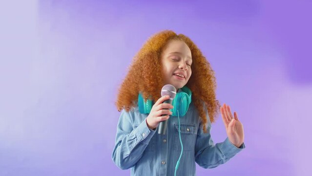 Studio Shot Of Girl Wearing Headphones Singing Karaoke Into Microphone Against Purple Background