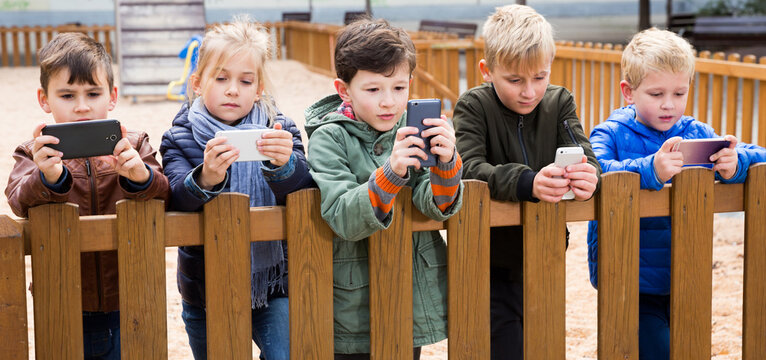Modern kids carried away with phone spending time together outdoors in autumn day