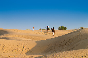 Fototapeta na wymiar Tourists riding camels, Camelus dromedarius, at sand dunes of Thar desert. Camel riding is a favourite activity amongst all tourists visiting here,
