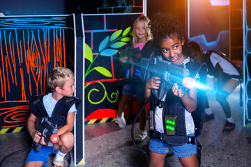 Happy african preteen girl with laser pistol playing laser tag with friends on dark labyrinth