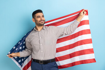 Man holding USA flag in hands, looking away with confident expression, proud of his social security in country, feeling in safe, wearing striped shirt. Indoor studio shot isolated on blue background.