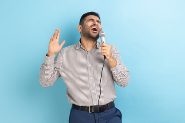 Fototapeta premium Portrait of attractive handsome young adult man with beard singing loud favorite songs with microphone in hands, wearing striped shirt. Indoor studio shot isolated on blue background.