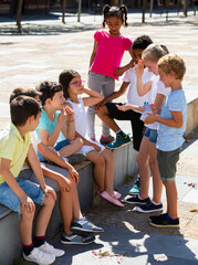 Group of cheerful children sitting on bench and sharing secrets outdoors
