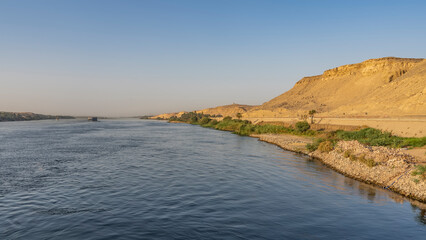 The wide blue river flows calmly. Ships are visible in the distance. There is green vegetation on the sandy shores. A high dune against a clear sky. Egypt. Nile