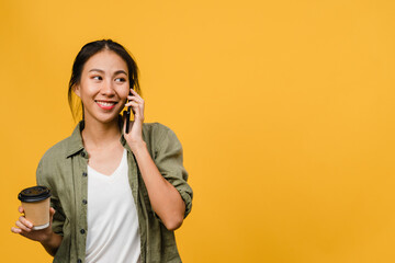 Young Asia lady talk by phone and hold coffee cup with positive expression, smile broadly, dressed in casual cloth feeling happiness and stand isolated on yellow background. Facial expression concept.