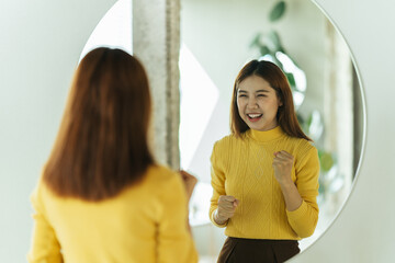 A young Asian woman talks to herself through a mirror to build her self-confidence and empower...