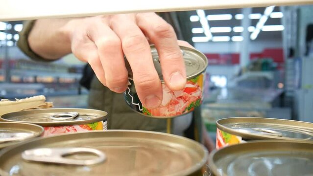 A man takes one can of precerved meat or fish on a supermarket shelf close-up
