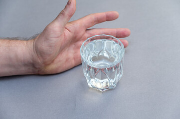 A man's hand intends to take a glass of water. A glass of clear drinking water against a gray background.