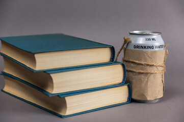 A stack of books and a can of drinking water against a gray background. Blue hardcover books. Can of freshwater wrapped in paper and tied with string.