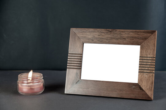 A wide brown empty photo frame set on a black surface next to a burning candle. Red Lighted candle in a clear glass jar. Horizontal placement of rectangular photo frame.