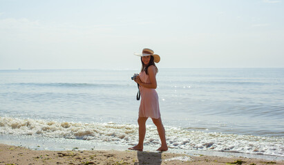 Happy young girl in a pink dress, straw hat and with digital camera going on the waves of the sea at noon. Relaxing unforgettable vacation at sea in summer. Enjoying summertime. Playing in water.