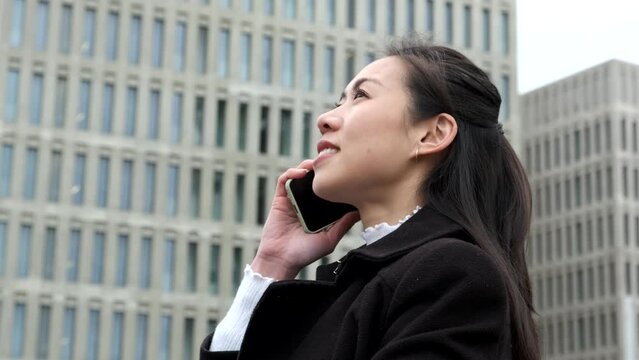 Low angle side view of happy Asian businesswoman in smart casual clothes. She is smiling and looking up while speaking on cellphone against skyscrapers on city street