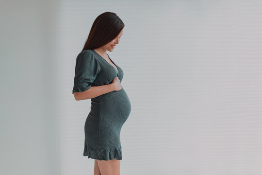 Beautiful pregnant young Asian woman looking down at her belly for maternity photoshoot wearing green dress. Pregnancy photos during third trimester