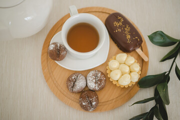 Coffee cookies, tartlet with berry jam and protein cream, ice cream in chocolate with a cup of tea on a wooden tray on a white tablecloth.