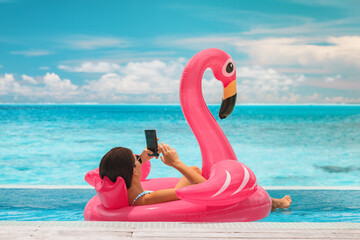 Summer swimming pool vacation relaxing woman floating in flamingo inflatable float using mobile phone at luxury resort sunbathing. Caribbean travel vacation hotel lifestyle - obrazy, fototapety, plakaty